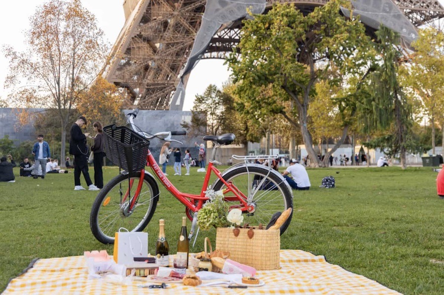 This is an image of a yellow, checkered picnic mat laid out with lots of artisan French food and wine. There is a bike and the Eiffel Tower in the background. This bike tour is a unique experience to try in Paris.
