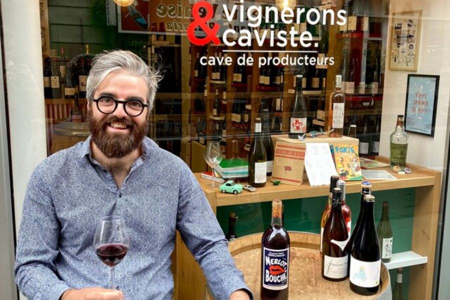 This is an image of a man sitting outside of a wine shop with a glass of red wine. He is smiling at the camera. On the table next to him are two other bottles of wine.